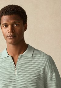 Light green zip-up polo shirt with a textured, honeycomb pattern and a ribbed collar. Features a silver zipper detail at the neck.