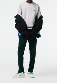 Sudadera gris de cuello redondo con un logo de Lacoste, chaqueta negra con acentos azules, pantalones deportivos verdes y zapatillas blancas con cordones verdes.