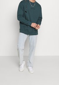 Felpa teal con dettagli rovinati e stampa "MENACE", abbinata a jeans blu chiaro e sneaker bianche. Design semplice, vestibilità casual.