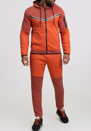 Survêtement en orange et rouge foncé. Sweat à capuche avec zip, poche avant et rayures contrastées. Jogging ajusté avec design bicolore. Accent noir.