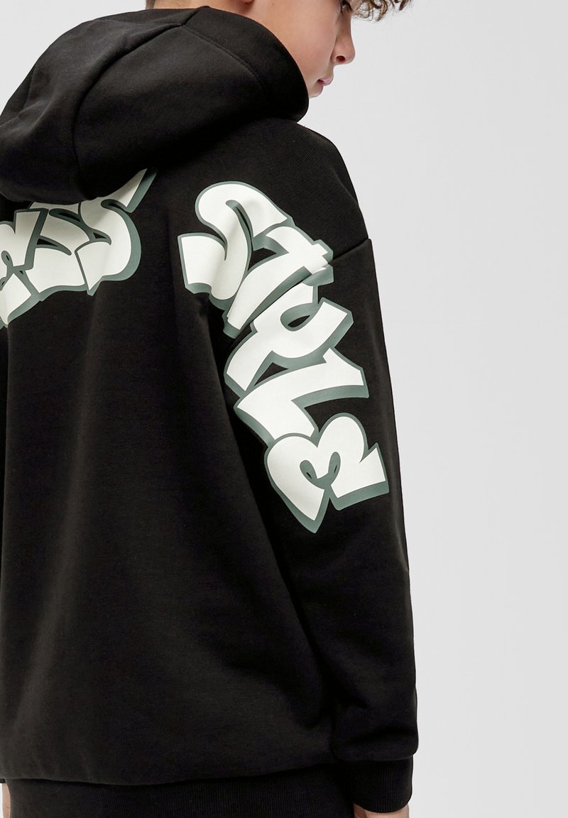 Zwarte hoodie met een gestructureerde afwerking, voorzien van oversized witte graffiti-achtige letters op de mouwen. Met capuchon en geribde manchetten.