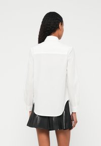 Balta krekla ar garām piedurknēm un pogu aizdari uz apkakles, ar taisnu griezumu un nedaudz garāku aizmugures malu. Valkāta kopā ar melnu krokotu svārku.