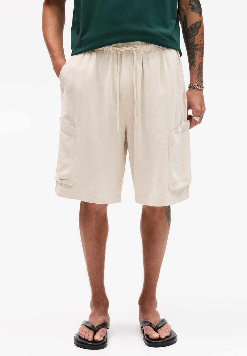 Shorts cargo beiges avec deux poches avant, une taille élastique et un cordon de serrage. Le tissu semble doux avec une légère texture. Tongs noires.