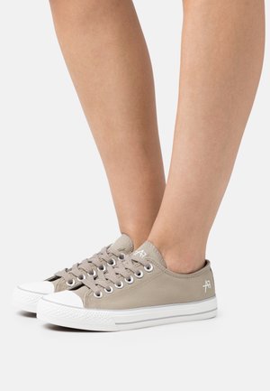 Dwa beżowe niskie trampki z płóciennego materiału z białymi gumowymi podeszwami i sznurowadłami, noszone na bose stopy na białym tle.