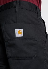 Pantalones de trabajo negros con una tela texturizada, con un bolsillo trasero y una etiqueta blanca con un logo amarillo. Diseñados para la durabilidad y la utilidad.