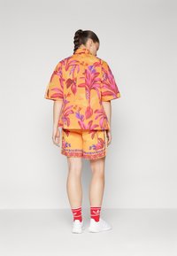 Oransje blomstrete kortermet skjorte med rosa og lilla mønstre, kombinert med oransje shorts med dekorativ kant. Hvite sneakers og røde sokker synlige.