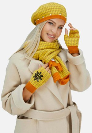 Frau trägt einen beigen Mantel mit passendem gelbem und orangefarbenem Hahnentritt-Hut, Schal und fingerlosen Handschuhen, wobei ein Handschuh ein schwarzes Schneeflocken-Design aufweist.