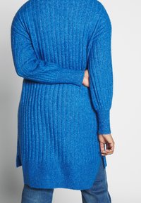 Grosse cardigan côtelée en bleu vif, avec un tissu texturé, des manches larges et des fentes sur les côtés, associé à un jean en denim.
