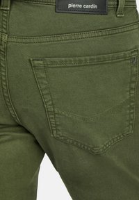 Pantalons en denim vert olive avec une poche arrière ornée d'un motif de couture en chevron, étiquette de marque et tissu texturé.