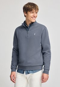 Grijze ritstrui van zachte stof, met een opstaande kraag, ribgebreide manchetten en een wit geborduurd logo op de linkerborst.