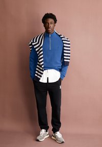 Sweatshirt zippé bleu superposé à une chemise blanche, jogging noir avec logo, et baskets grises. Écharpe rayée horizontale drapée sur les épaules.