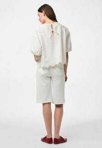 Vit blus med korta ärmar som har en scallopad nederkant och ögla detaljer längst ner, matchad med vita shorts och röda skor.