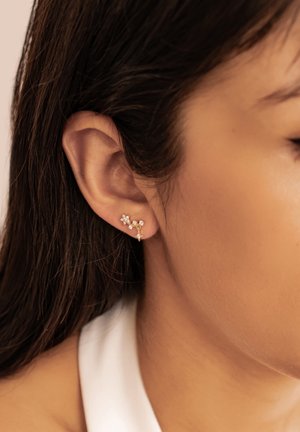 Gold ear cuff earring with small clear gemstones worn on the upper earlobe of a person with straight dark hair and light brown skin.