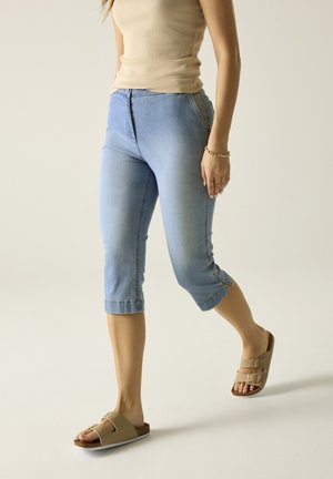 Vrouw draagt een beige tanktop, lichtblauwe korte spijkerbroek, beige sandalen met gesp en een gouden armband, lopend tegen een effen achtergrond.