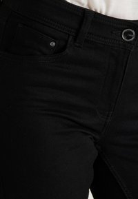 Jeans de mezclilla negra con un corte ajustado, que presentan un pequeño bolsillo delantero, herrajes de metal y detalles de costura en contraste. Textura suave.