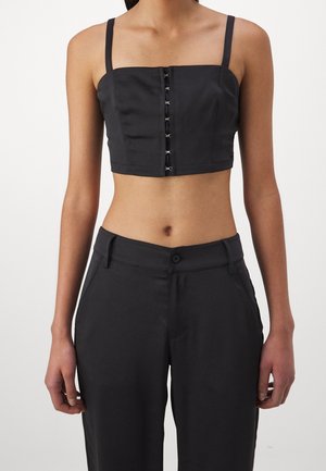 Kobieta ubrana w czarny krótki top z przednimi zapięciami na haftki oraz czarne, wysokie spodnie na tle jednolitego tła.