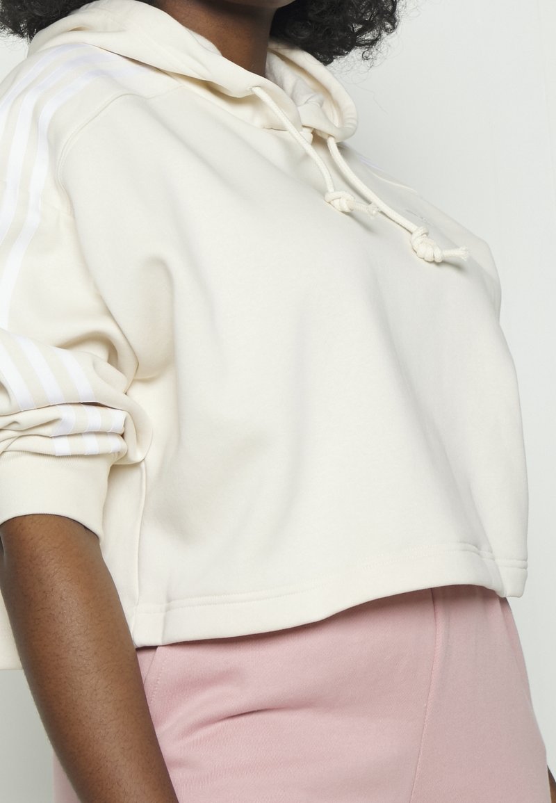 Krátká krémová mikina s kapucí a stahovací šňůrkou, s bílými proužky na rukávech. Měkká látka s hladkou texturou. Doplněná světle růžovými kalhotami.