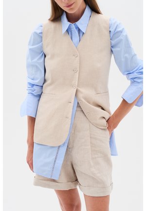 Vrouw draagt beige knoopvest over blauw overhemd met lange mouwen en beige opgerolde korte broek, hand in zak tegen effen achtergrond.