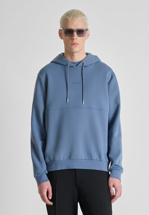 Persona joven con cabello corto y rubio, que lleva gafas de sol grandes y cuadradas, una sudadera azul con capucha y pantalones negros sobre un fondo liso.