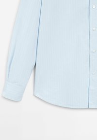 Light blue long-sleeve shirt with vertical white stripes, button closure, and a curved hem. Made from smooth fabric with a structured fit.