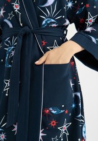 Mörkblå morgonrock med ett himmelskt mönster som har stjärnor och månar. Mjukt tyg, vida ärmar och ett bälte i midjan. Inkluderar en sidoficka.