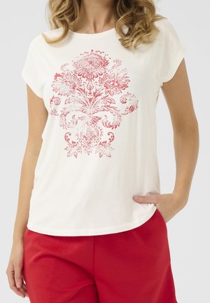 Kobieta ubrane w białą koszulkę z krótkim rękawem z czerwonym kwiatowym wzorem damask i czerwone spodnie, jedna ręka w kieszeni, widoczny tułów.