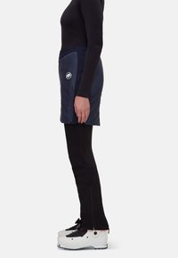 Marinsky isolerad kjol med quiltad design, sidodragkedja och logodetalj, bärs över svarta tajta byxor och vita skidstövlar.