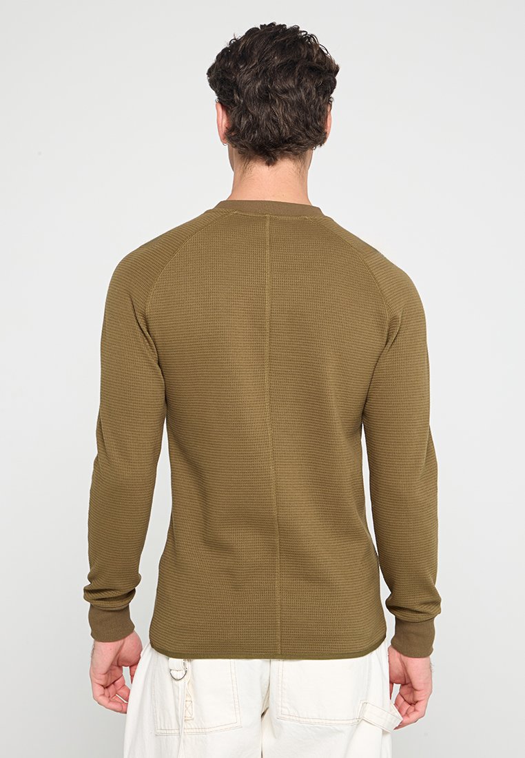 Mann trägt ein olivgrünes, strukturiertes Langarmshirt und off-white Hosen, steht mit dem Rücken zu einem schlichten Hintergrund.