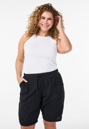 Lachende vrouw met krullend haar, gekleed in een wit mouwloos topje en zwarte shorts met trekkoord, poserend met één hand in de zak en één hand die haar haar aanraakt.