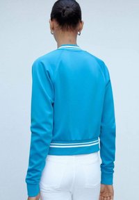 Giacca bomber blu brillante con zip frontale, polsini e vita a coste con righe bianche, caratterizzata da un tessuto liscio e strutturato.