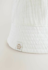White cotton bucket hat with textured fabric, featuring a sewn label that reads “hello little one” in muted gold lettering.