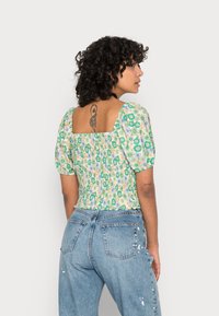 Blusa floral verde com mangas bufantes e costas quadradas, feita de tecido leve. Combinada com jeans de denim azul com detalhes desgastados.