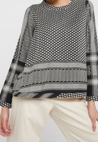 Maglietta a maniche lunghe bianca e nera con colletto rotondo, caratterizzata da disegni geometrici e accenti testurizzati. Indossata sopra pantaloni chiari.