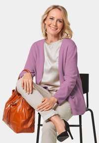 Lila Strickjacke, leichtes Seidentop, cremefarbene Hose und schwarze High Heels. Braune Lederhandtasche mit Kordelzugdetails. Minimalistische Umgebung.