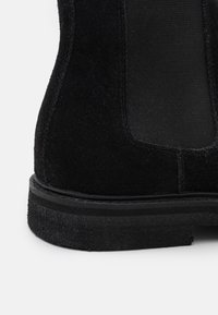 Botín negro de ante con paneles laterales elásticos, suela plana de goma y un acabado texturizado. Presenta un diseño elegante y minimalista.