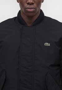 Gros plan d'une personne portant une veste noire avec une fermeture éclair et un petit logo de crocodile vert sur la poitrine.