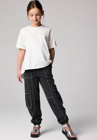 Valkoinen lyhythihainen t-paita, jossa on teksturoitu kuvio, yhdistettynä mustiin cargo-housuihin, joissa on taskuja ja valkoiset tikkaukset, sekä metallinvärisiin sandaaleihin.