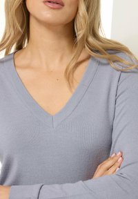 Lichtgrijze V-hals trui met geribbelde halslijn en lange mouwen. Zachte textuur en getailleerde pasvorm, casual gedragen met de armen gekruist.