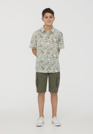 Camicia a maniche corte con bottoni di un verde chiaro e un motivo a foglia bianca, abbinata a pantaloni cargo verde oliva e sneakers bianche.