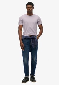 T-shirt lilas à manches courtes, jeans en denim indigo, sweat-shirt bleu marine noué autour de la taille, chaussures noires à lacets, pose debout.