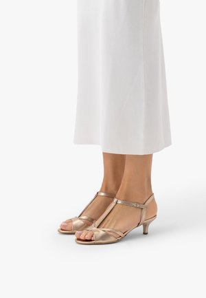 Stopy w metalicznie różowo-złotych sandałach na niskim obcasie z paskiem w kształcie litery T i odkrytymi palcami, zestawione z białą spódnicą sięgającą kostek.