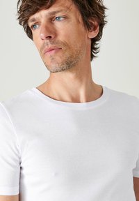 T-shirt bianco a maniche corte con collo rotondo, realizzato in tessuto liscio e traspirante. Presenta un design semplice e aderente senza fantasie.