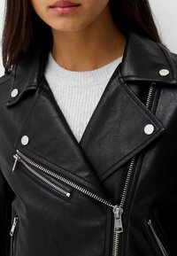 Czarna kurtka motocyklowa z sztucznej skóry z srebrnym osprzętem, z zamykanymi kieszeniami i charakterystycznym kołnierzem, noszona na jasnoszarym swetrze.