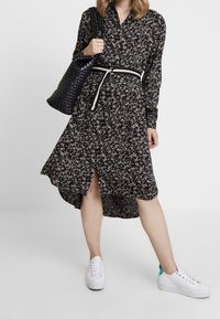 Czarna sukienka w kwiaty z długimi rękawami, wysokim kołnierzem i asymetrycznym dołem. Zestawiona z czarną plecioną torbą i białymi trampkami. Posiada biały pasek.