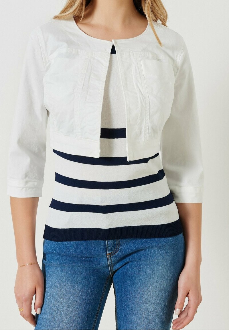Femme portant une veste blanche courte à manches trois-quarts sur un haut rayé blanc et marine associée à un jean bleu.