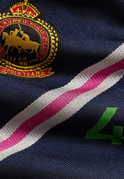 Υφασμα ναυτικού μπλε με κόκκινη, λευκή και ροζ ρίγα, πράσινο αριθμό 4 και κεντημένο χρυσό έμβλημα που δείχνει δύο παίκτες πόλο κάτω από ένα στέμμα.