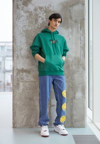 Felpa verde con tasca frontale e logo, jeans di denim blu con grafiche di faccine sorridenti gialle, abbinati a sneakers bianche e rosse.