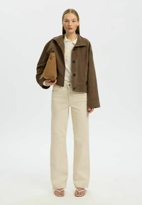 Jeune femme portant une veste courte marron, un polo crème, un pantalon large crème, des sandales beiges, tenant un sac à main en daim marron sur fond blanc.