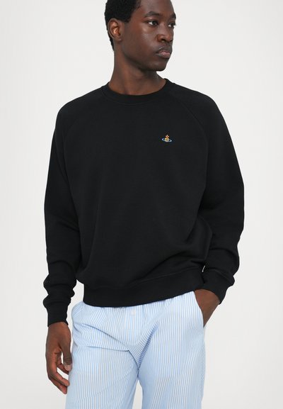 Schwarzer Pullover mit Raglanärmeln, auf dessen Brust sich ein kleines, buntes Logo befindet. Der Stoff wirkt glatt und bequem. Gestreifte hellblaue Hose.