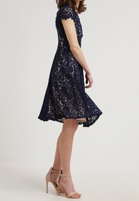 Tmavě modré krajkové šaty s krátkými zvlněnými rukávy a rozevlátým lemem, doplněné oholenými vysokými podpatky, které předvádějí složité krajkové vzory.